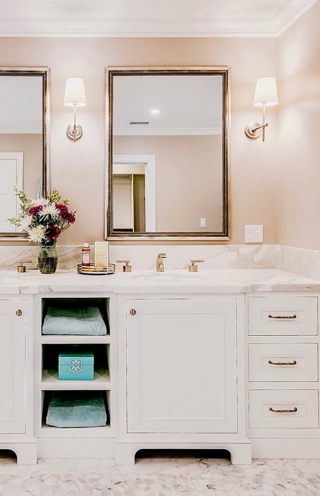 Primary bath storage solutions presented in a beautiful Ggem Design luxury bathroom with cream cabinets and peach walls sconces and high end mirrors and cabinet hardware